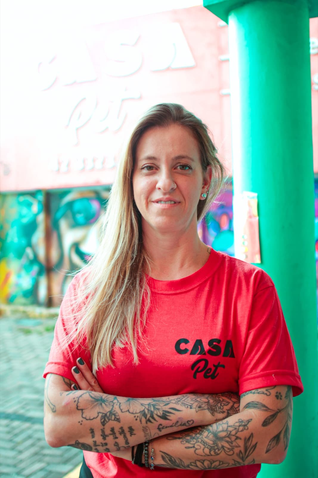 Retrato da Tia Drê encostada em uma coluna verde da creche, com o mural ao fundo