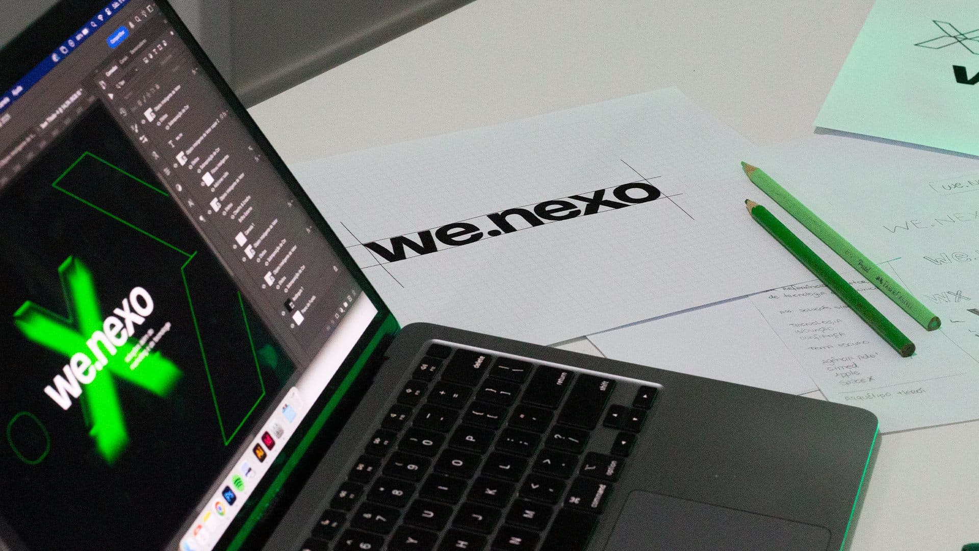 Bastidor do ensaio fotográfico da We.nexo: macbook exibindo o logo verde, papel quadriculado com construção do logotipo e lápis sobre a mesa